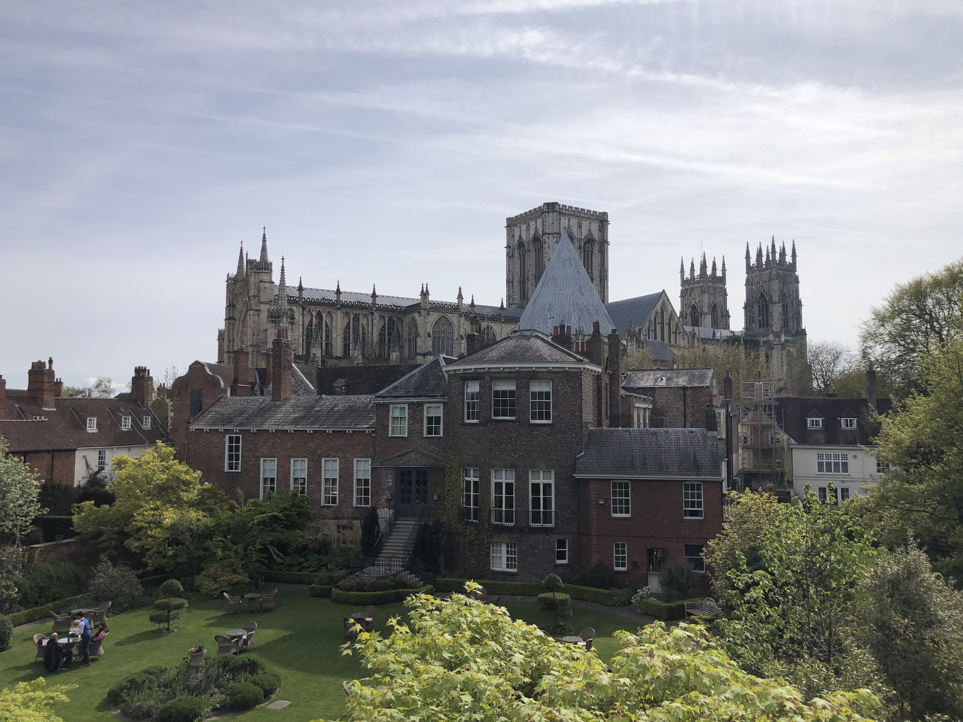 The York Minster is seen in the distance, the sky is blue with wispy clouds. Buildings obscure the lower half of the Minster.