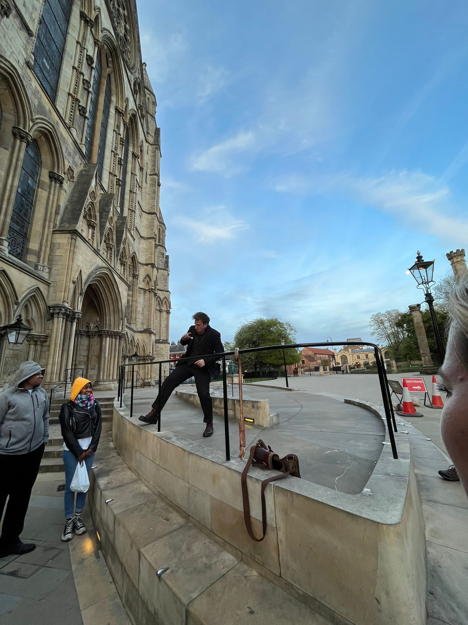 Dorian hunched over as he tells a ghost story, along side the York Minster, the evening sky in the background bright blue.